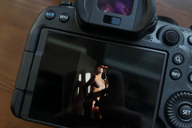 Aparat fotograficzny leży na drewnianej podłodze. Wyświetlacz aparatu przedstawia zdjęcie na którym jest sesja zdjęciowa 2026. Młoda kobieta pozuje do zdjęcia w bieliźnie. Dziewczyna ma na sobie jasny pudroworóżowy biustonosz 